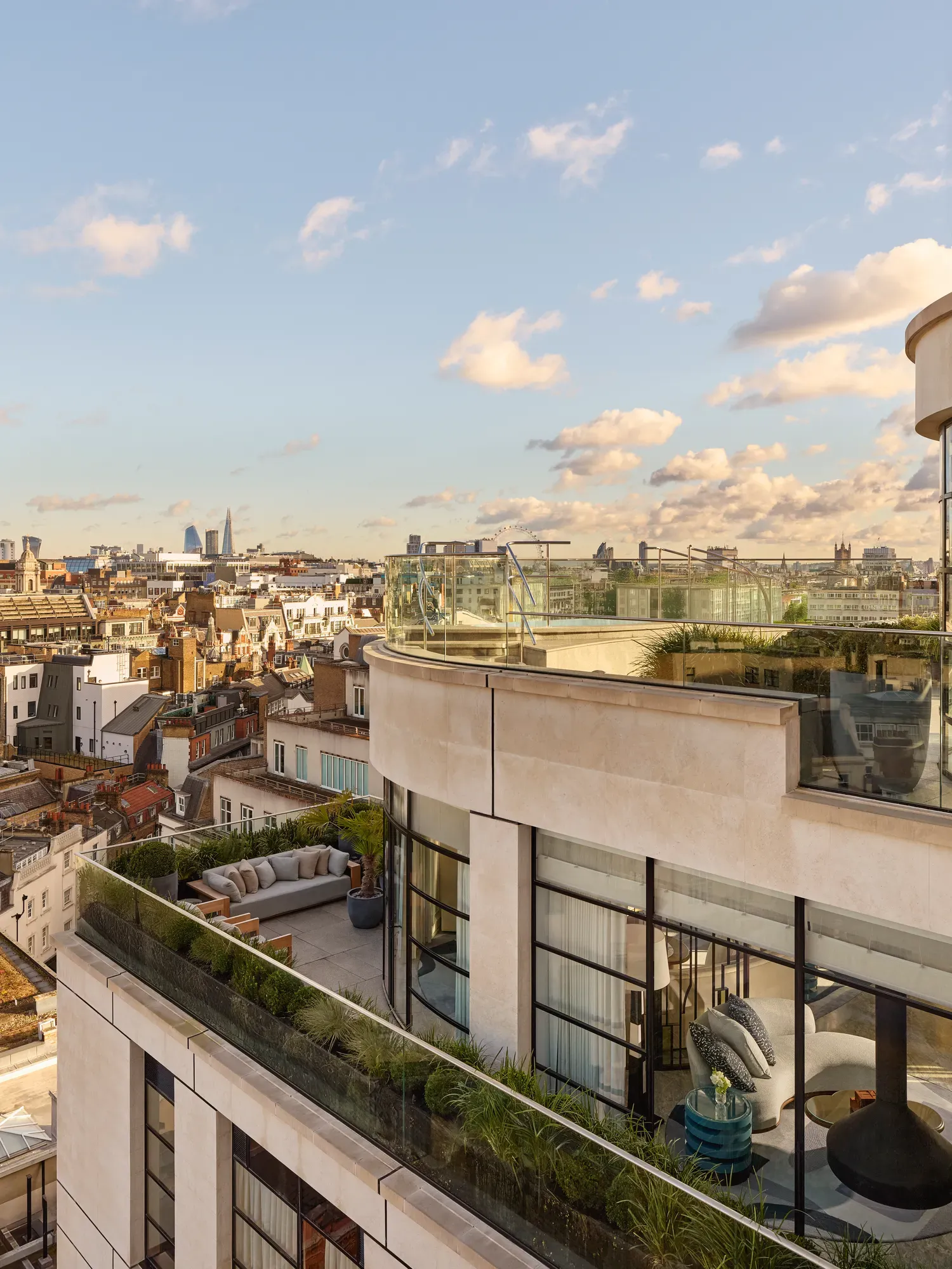 Exterior view of a luxury penthouse with wraparound terraces featuring outdoor seating, greenery, and glass balustrades, overlooking London’s skyline under a clear evening sky.