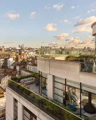 Exterior view of a luxury penthouse with wraparound terraces featuring outdoor seating, greenery, and glass balustrades, overlooking London’s skyline under a clear evening sky.