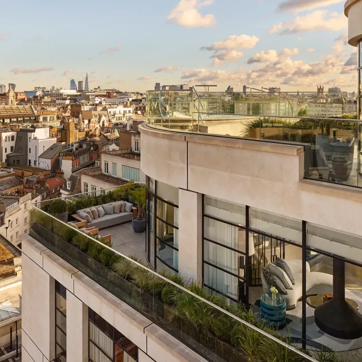 Exterior view of a luxury penthouse with wraparound terraces featuring outdoor seating, greenery, and glass balustrades, overlooking London’s skyline under a clear evening sky.