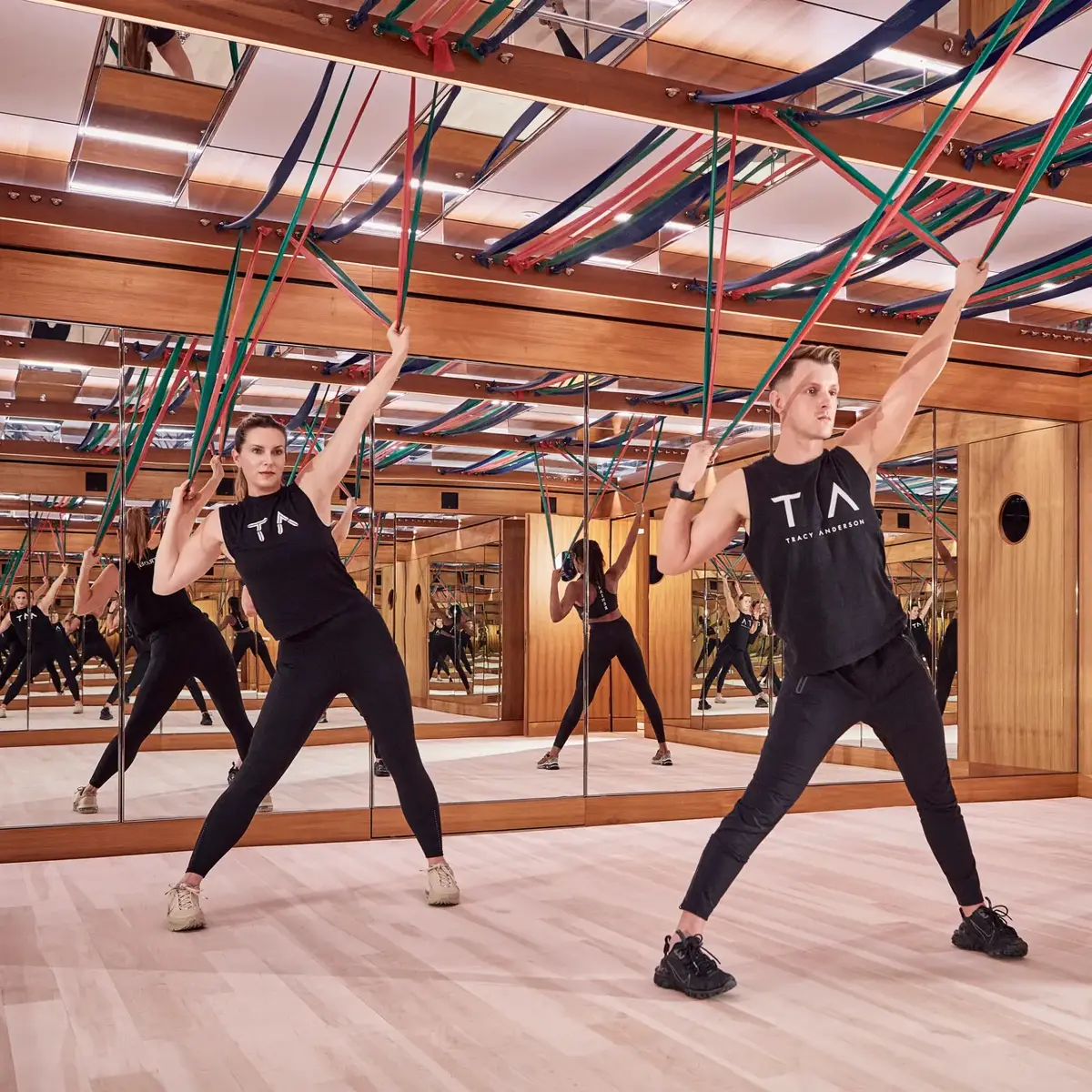 Group fitness class using resistance bands in a mirrored studio with wood panelling and suspended straps.