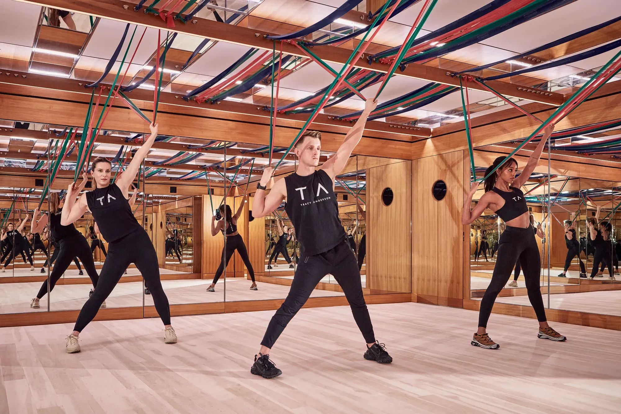 Group fitness class using resistance bands in a mirrored studio with wood panelling and suspended straps.