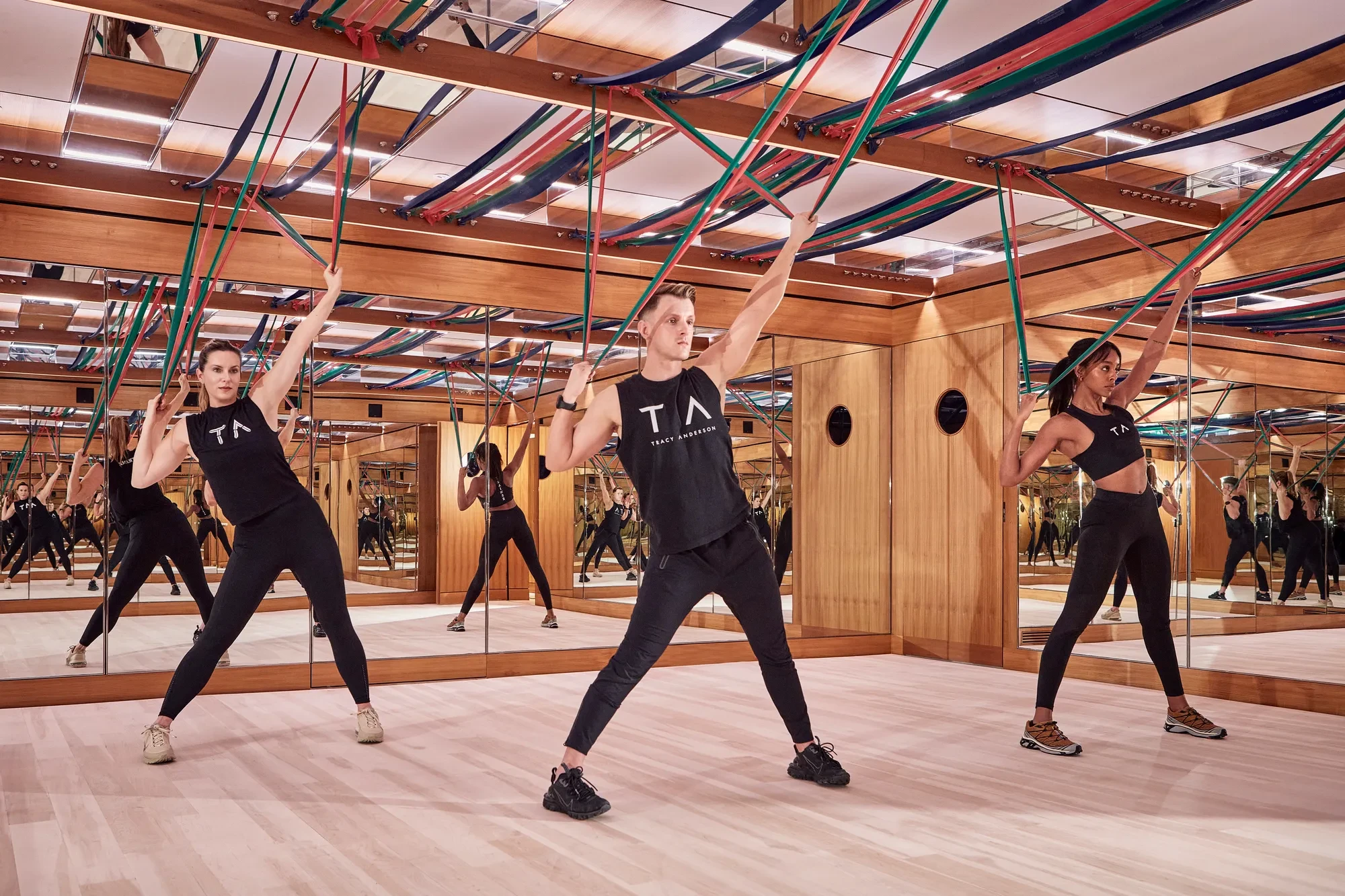 Group fitness class using resistance bands in a mirrored studio with wood panelling and suspended straps.