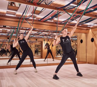 Group fitness class using resistance bands in a mirrored studio with wood panelling and suspended straps.