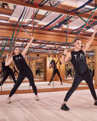 Group fitness class using resistance bands in a mirrored studio with wood panelling and suspended straps.