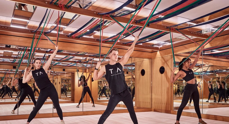 Group fitness class using resistance bands in a mirrored studio with wood panelling and suspended straps.
