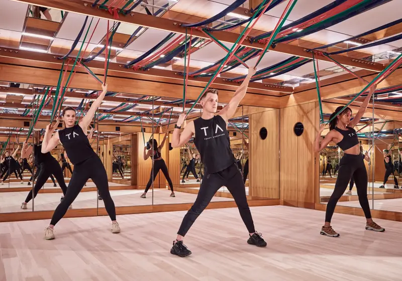 Group fitness class using resistance bands in a mirrored studio with wood panelling and suspended straps.