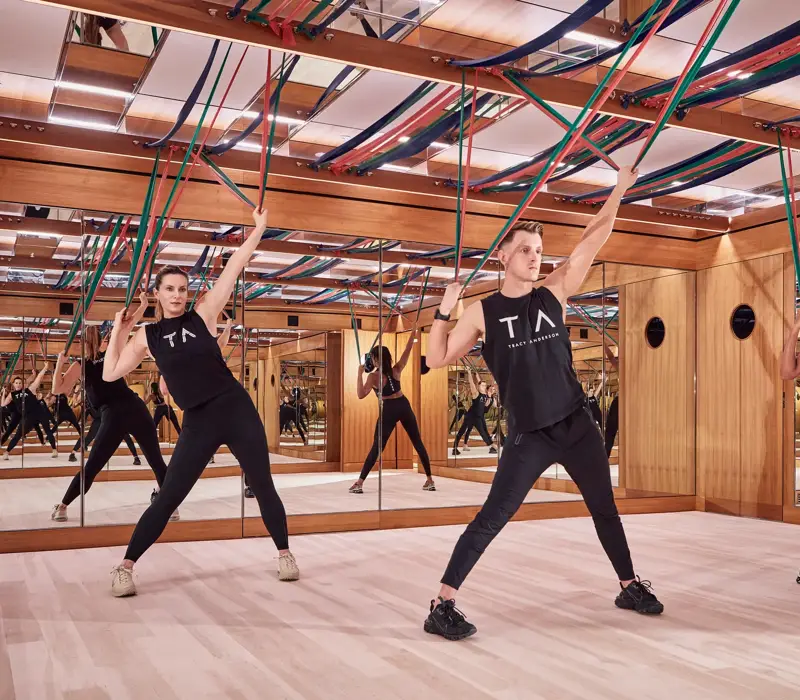 Group fitness class using resistance bands in a mirrored studio with wood panelling and suspended straps.
