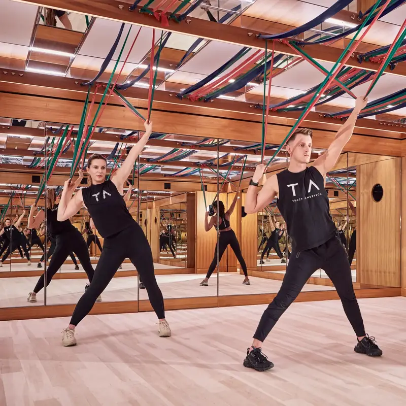 Group fitness class using resistance bands in a mirrored studio with wood panelling and suspended straps.