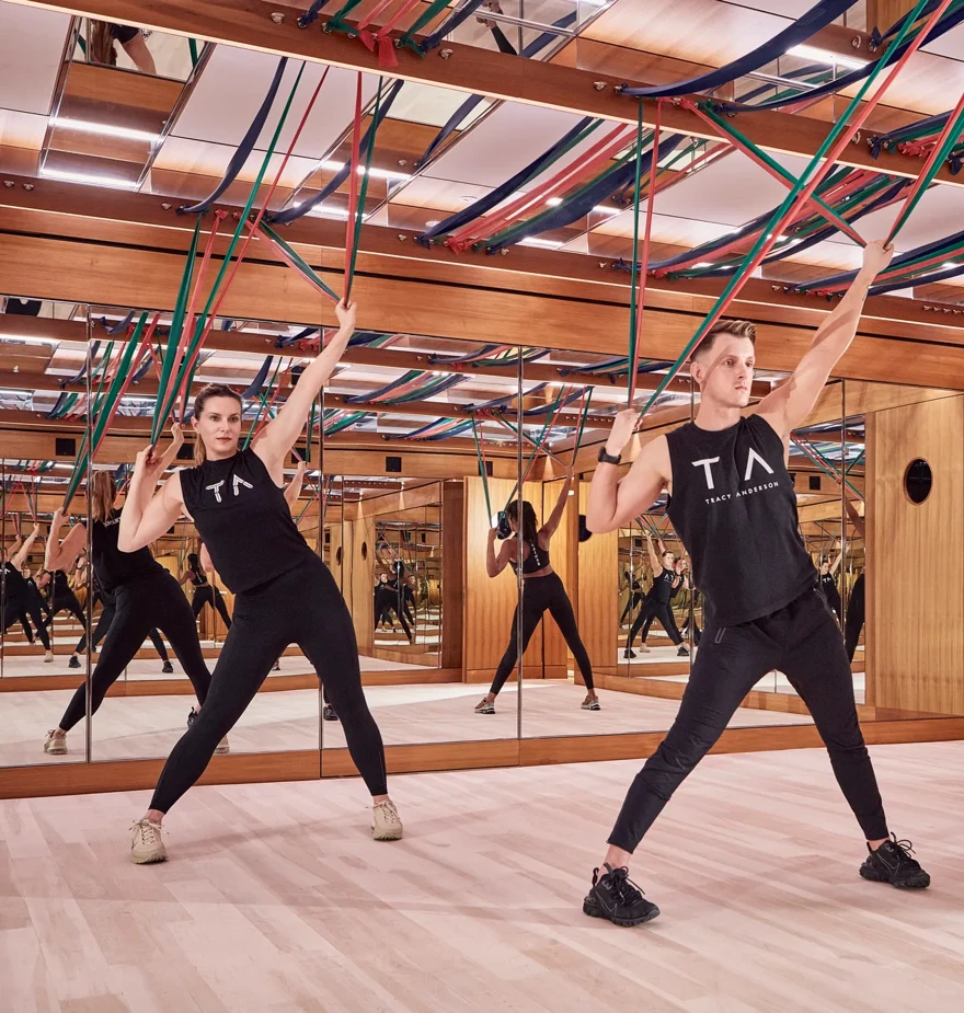 Group fitness class using resistance bands in a mirrored studio with wood panelling and suspended straps.