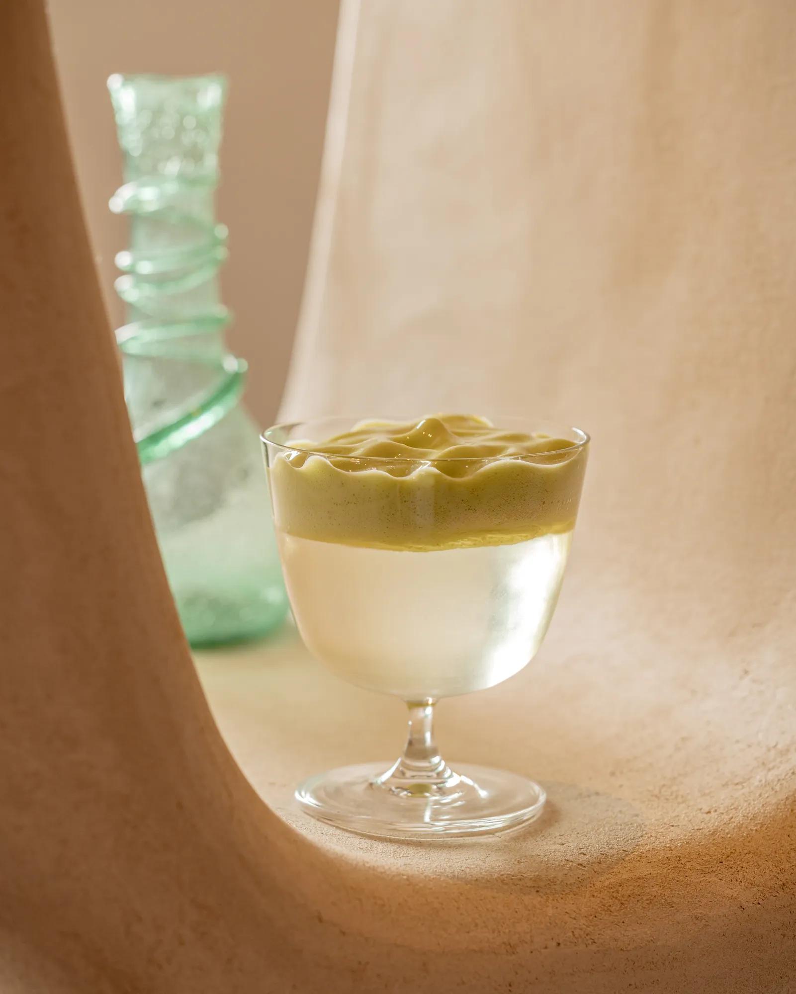 Coupe glass filled with a pale cocktail topped with a textured foam layer, displayed in a sculptural alcove beside a green glass bottle.