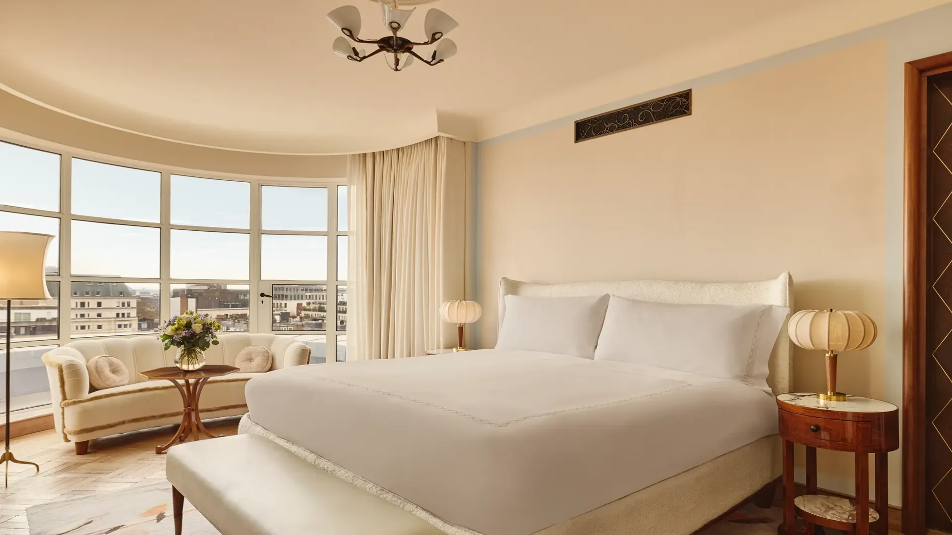 Bright bedroom with a curved window overlooking London, featuring a king-size bed, cream sofa with round cushions, and warm neutral decor with vintage-style lighting.