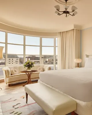 Bright bedroom with a curved window overlooking London, featuring a king-size bed, cream sofa with round cushions, and warm neutral decor with vintage-style lighting.