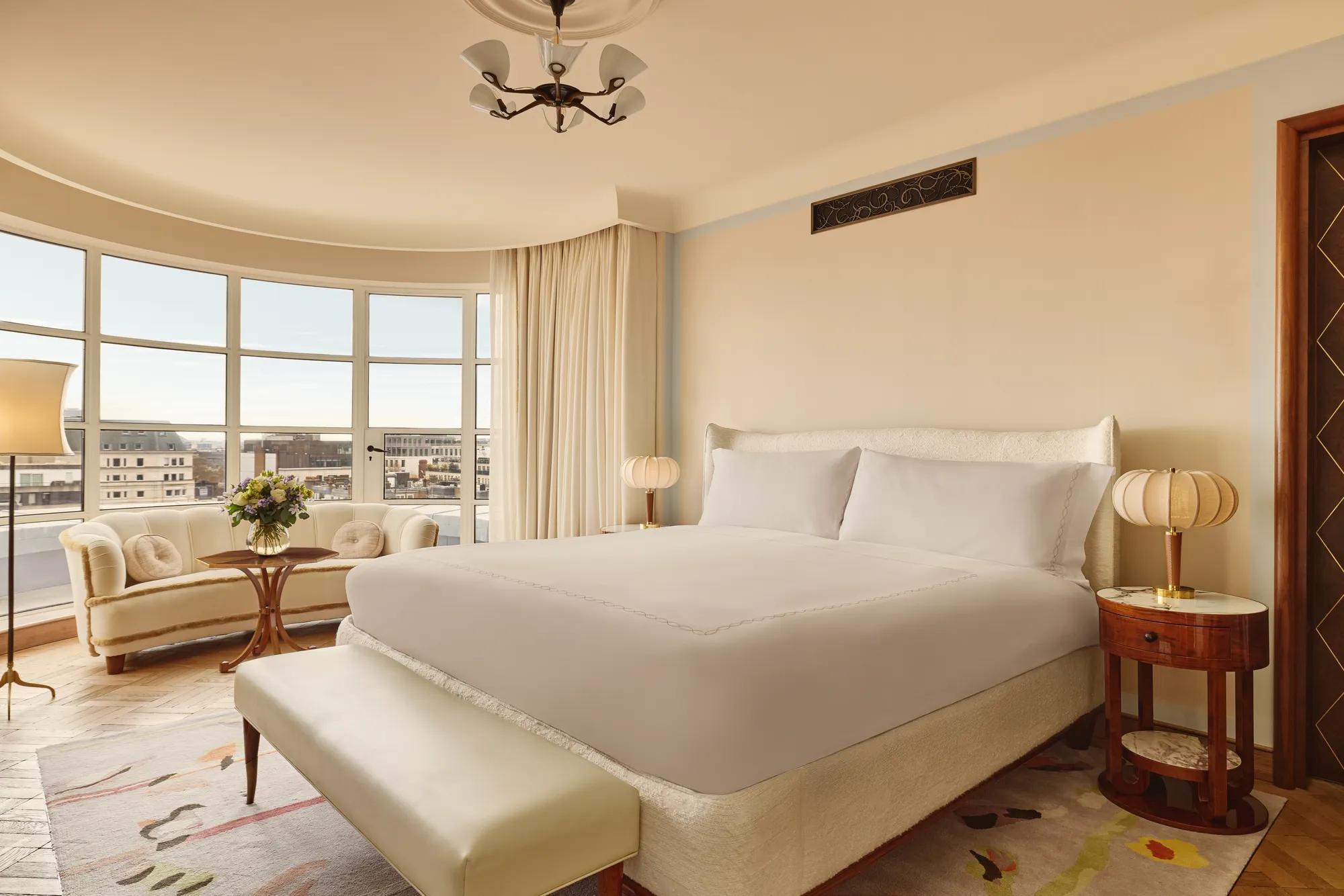 Bright bedroom with a curved window overlooking London, featuring a king-size bed, cream sofa with round cushions, and warm neutral decor with vintage-style lighting.