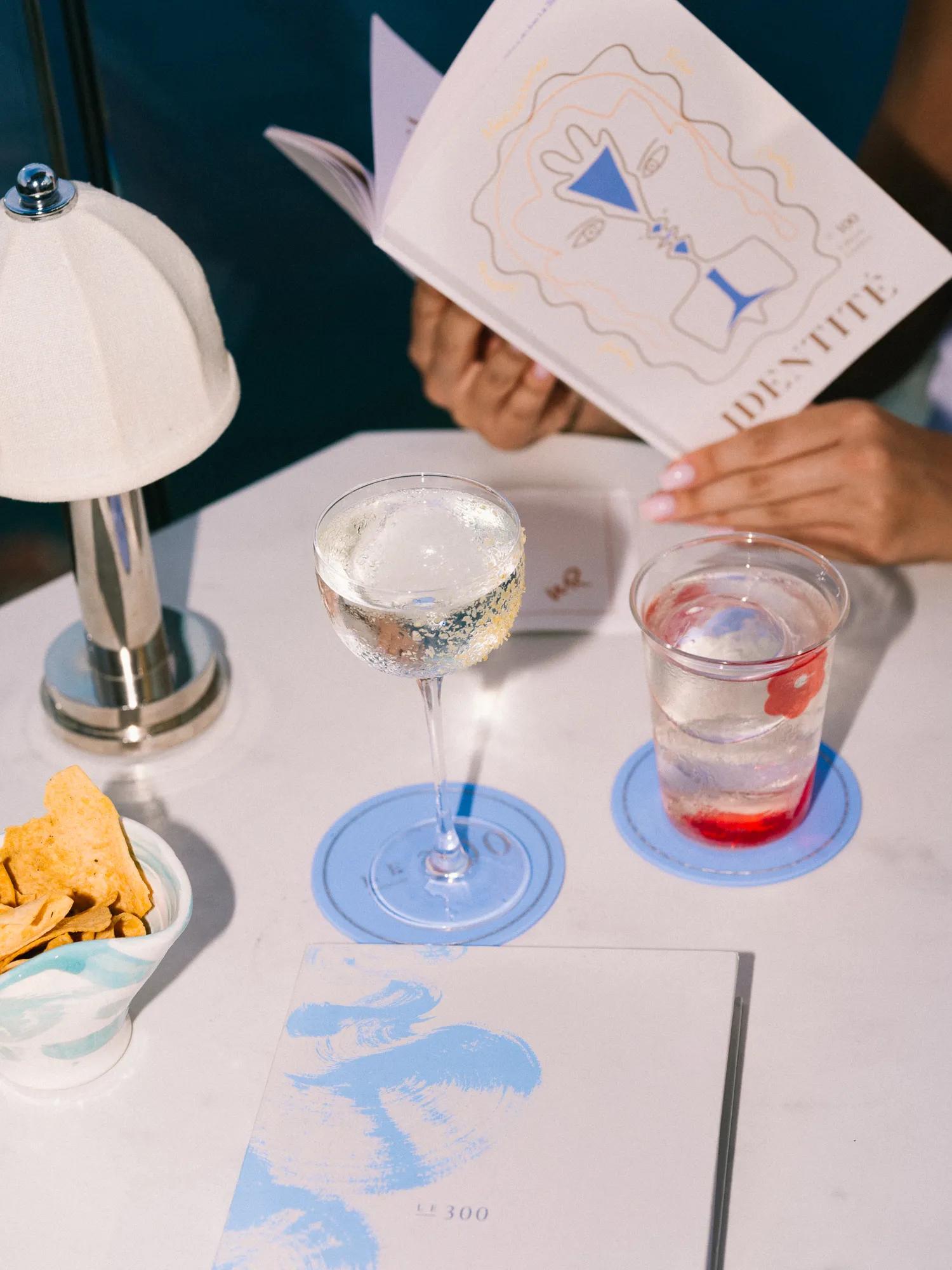 Une personne lit un menu de cocktails à une table extérieure avec des boissons, des en-cas et une petite lampe.
