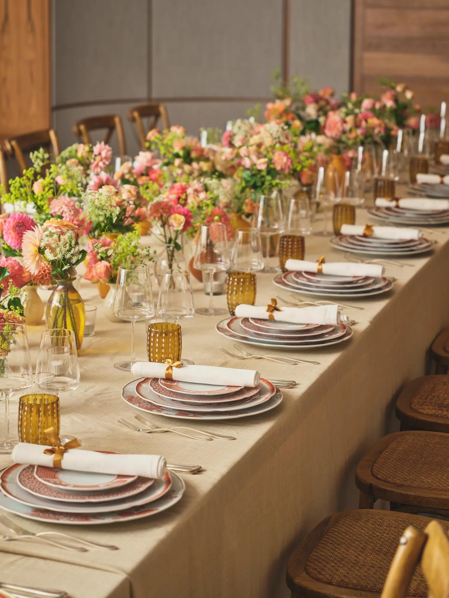 Long dining table with layered plates, amber glasses, and vibrant floral arrangements.