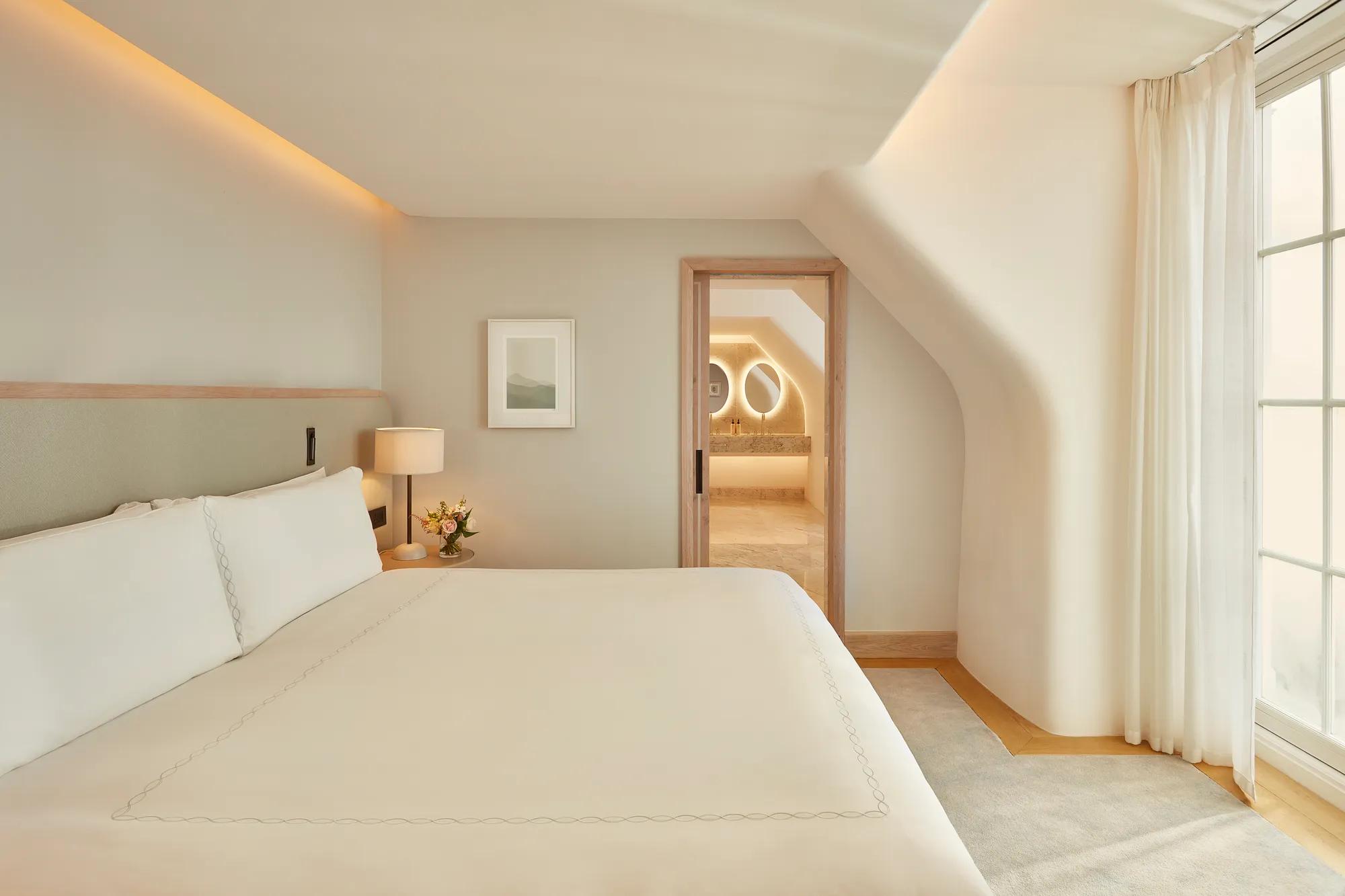 Serene bedroom with a large bed, soft neutral tones, and natural light from a window, leading to a marble bathroom with illuminated mirrors through an open doorway.