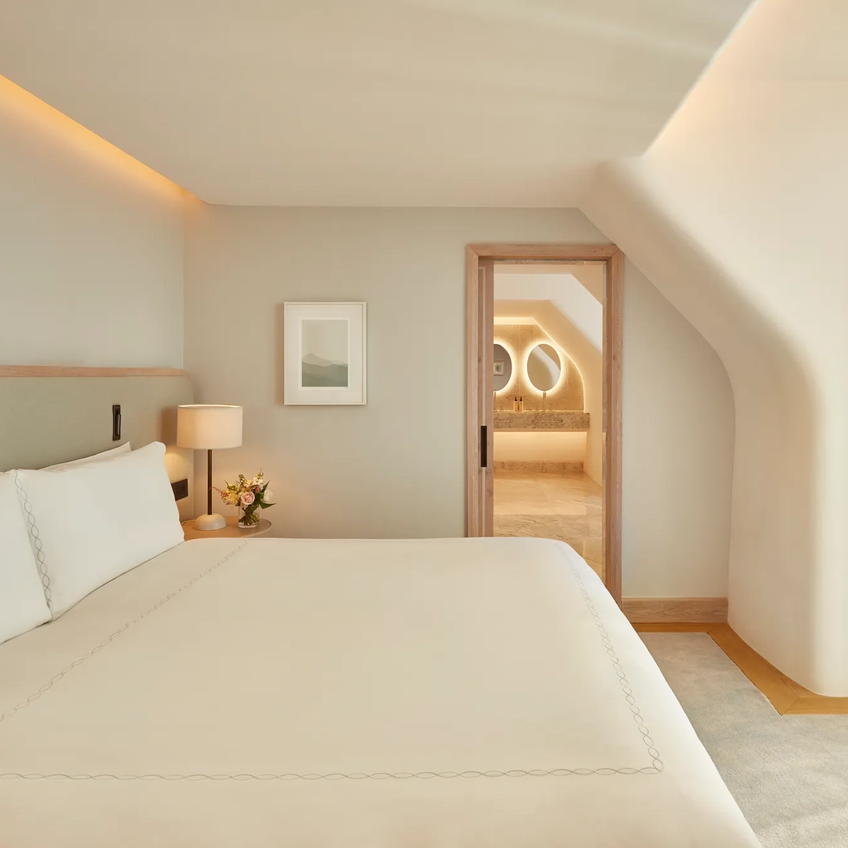 Serene bedroom with a large bed, soft neutral tones, and natural light from a window, leading to a marble bathroom with illuminated mirrors through an open doorway.