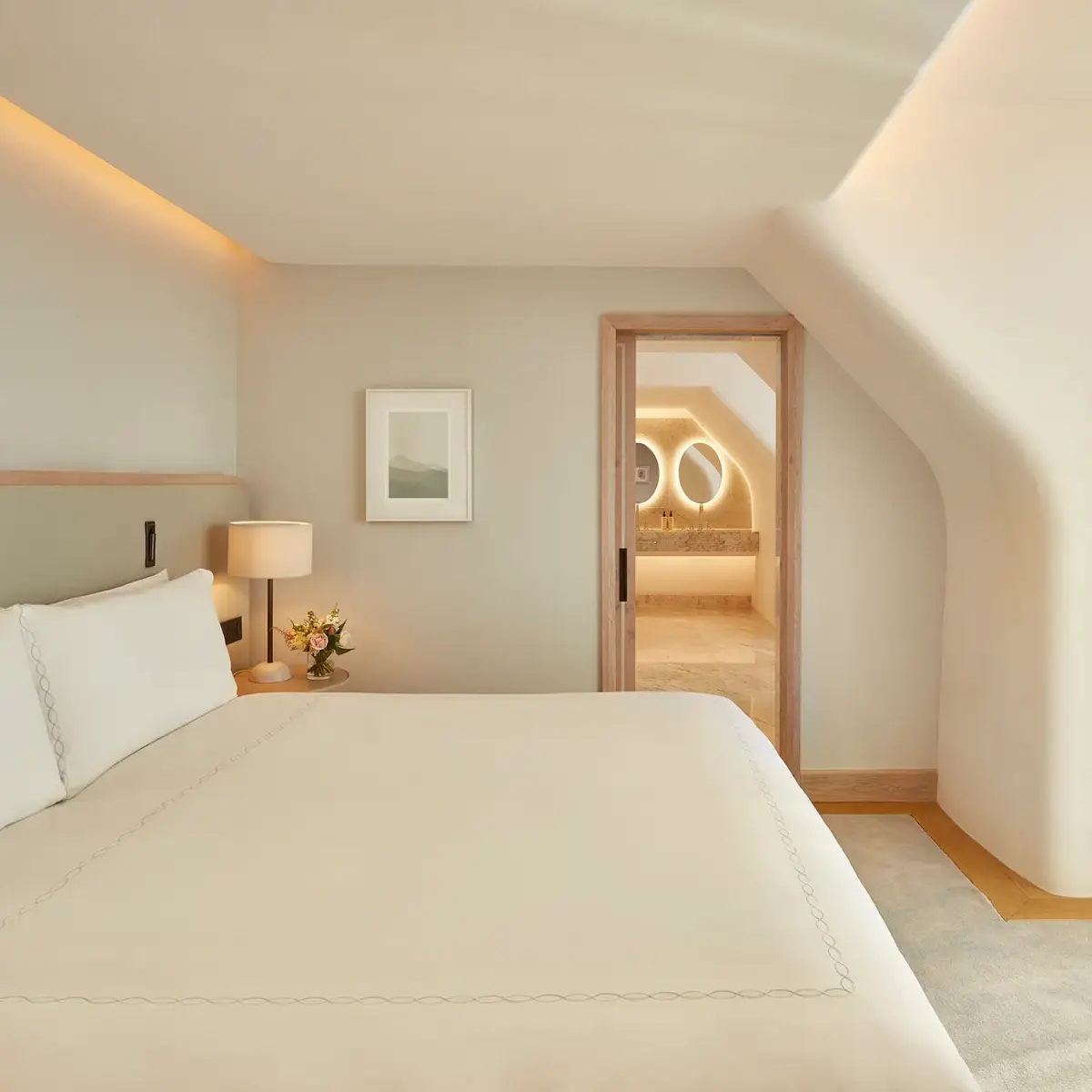 Serene bedroom with a large bed, soft neutral tones, and natural light from a window, leading to a marble bathroom with illuminated mirrors through an open doorway.