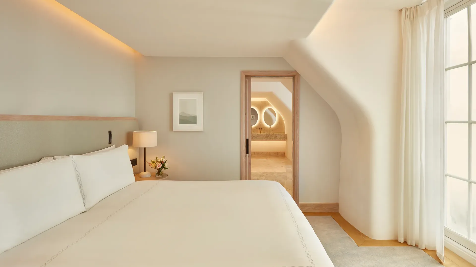 Serene bedroom with a large bed, soft neutral tones, and natural light from a window, leading to a marble bathroom with illuminated mirrors through an open doorway.