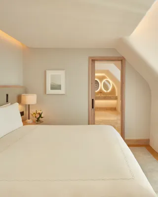 Serene bedroom with a large bed, soft neutral tones, and natural light from a window, leading to a marble bathroom with illuminated mirrors through an open doorway.