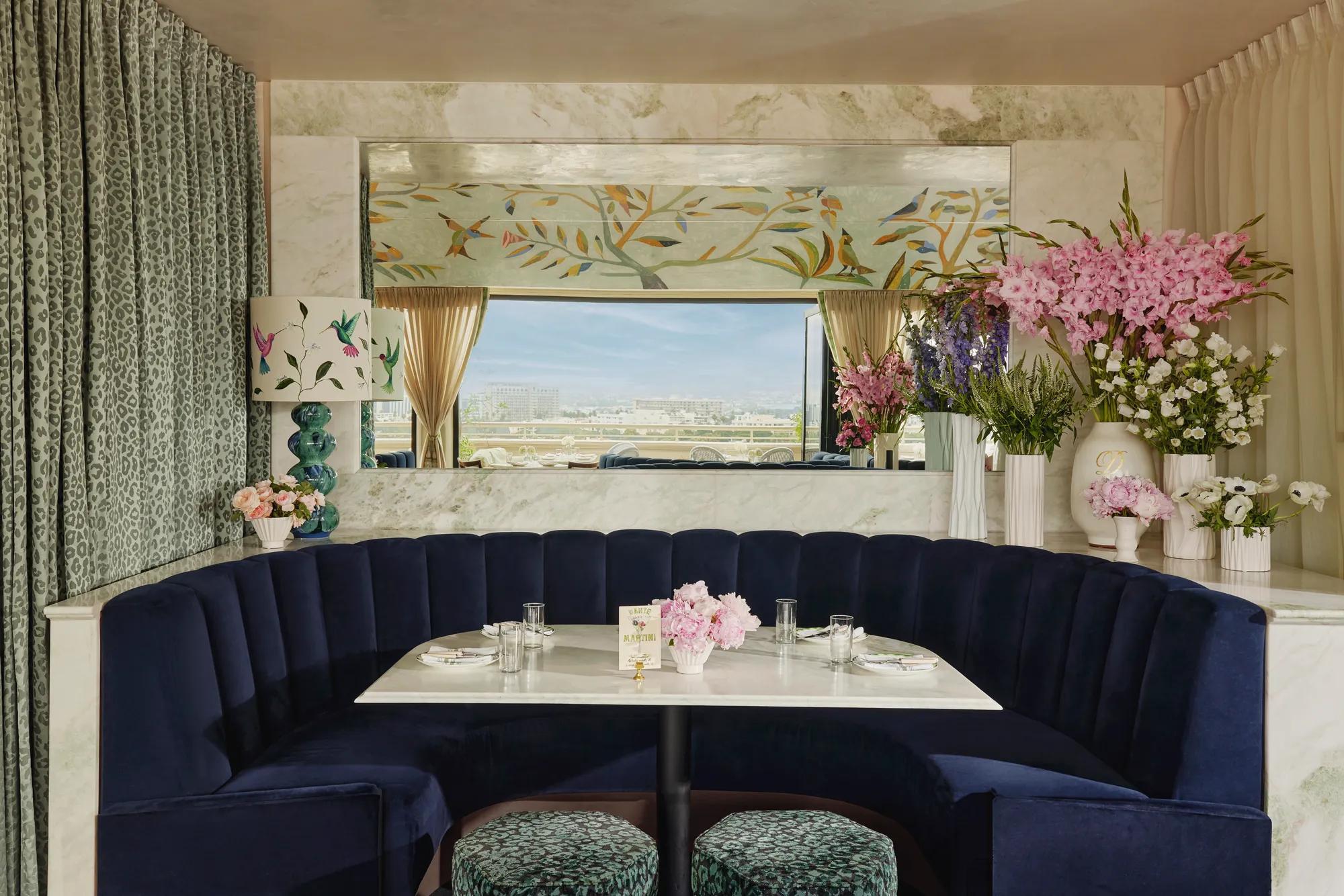Intimate interior blue-hued booth with semicircular navy banquette, marble table, and abundant flowers; mirror reflects the open-air terrace beyond.