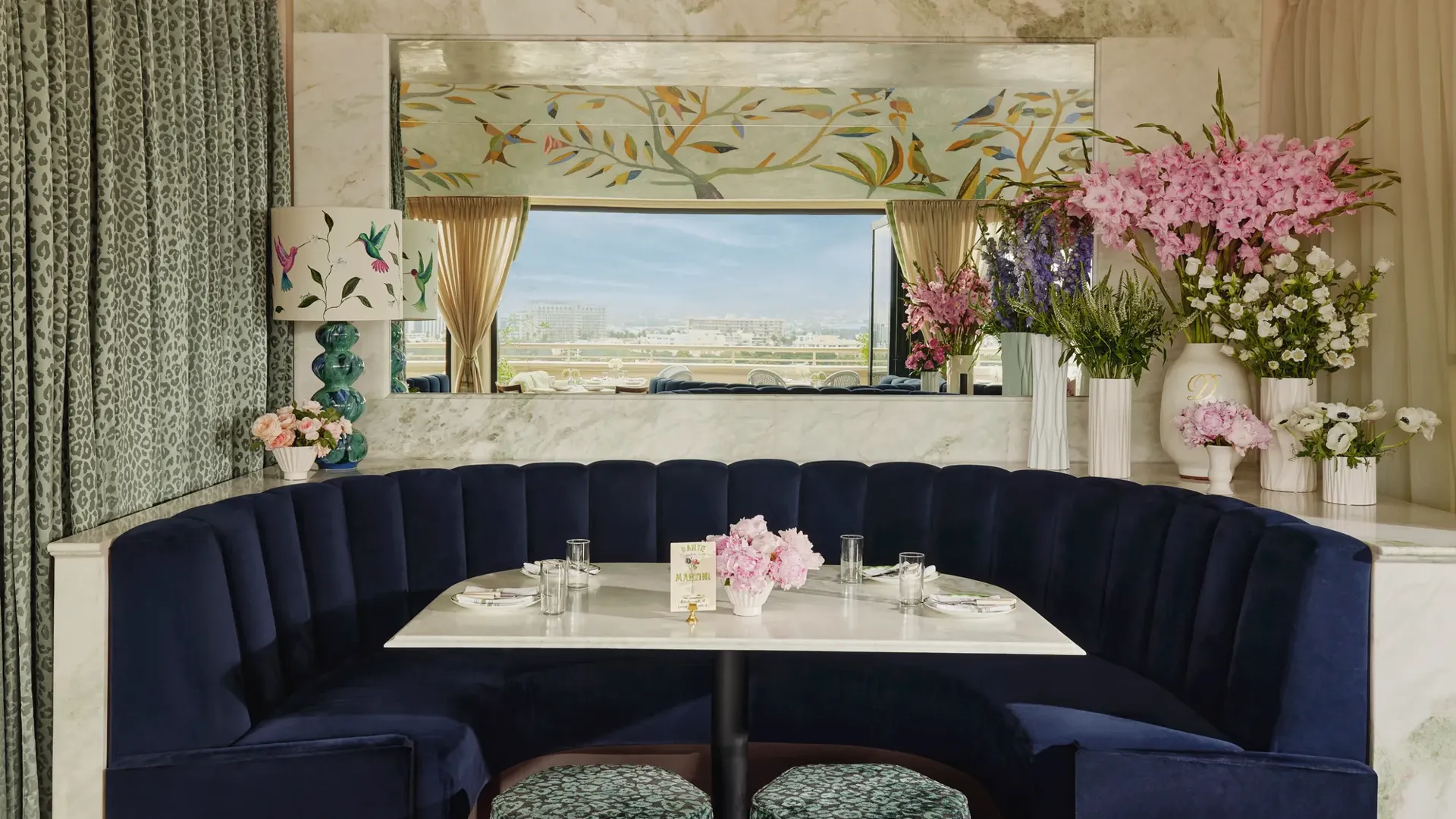 Intimate interior blue-hued booth with semicircular navy banquette, marble table, and abundant flowers; mirror reflects the open-air terrace beyond.