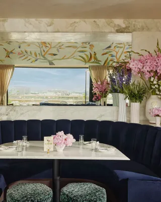 Intimate interior blue-hued booth with semicircular navy banquette, marble table, and abundant flowers; mirror reflects the open-air terrace beyond.
