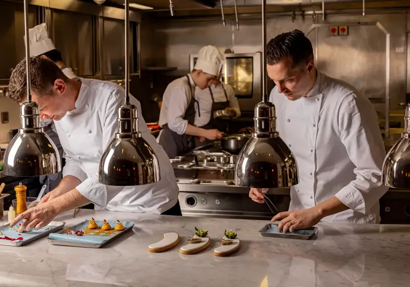 Chefs in a professional kitchen carefully plating dishes under heat lamps.