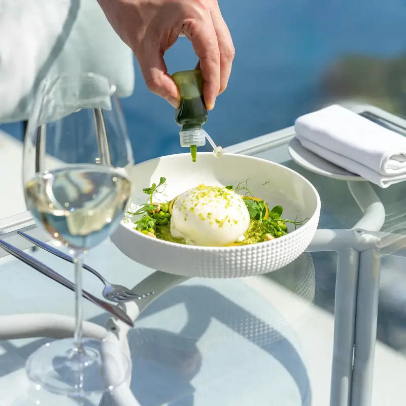 A hand pours green herb oil over a burrata dish on a terrace table overlooking the sea.