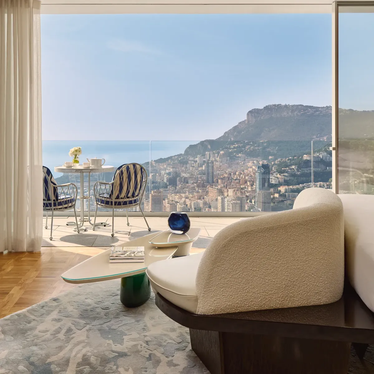 Chambre d’hôtel luxueuse avec fauteuil moderne et table basse, s’ouvrant sur une terrasse avec chaises rayées et vue sur la ville et la mer.