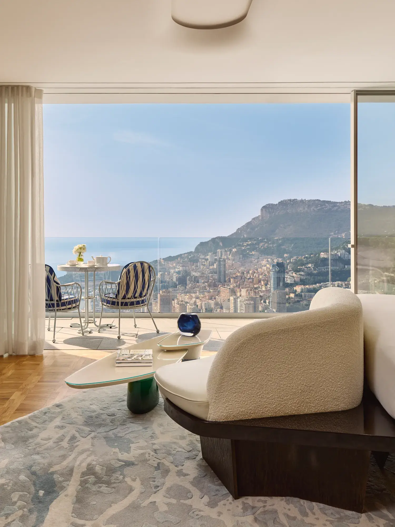 Chambre d’hôtel luxueuse avec fauteuil moderne et table basse, s’ouvrant sur une terrasse avec chaises rayées et vue sur la ville et la mer.