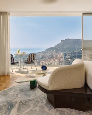 Chambre d’hôtel luxueuse avec fauteuil moderne et table basse, s’ouvrant sur une terrasse avec chaises rayées et vue sur la ville et la mer.