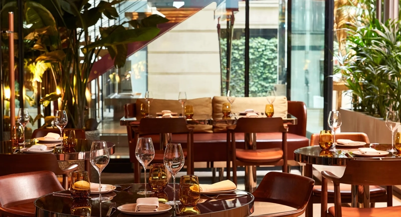 A contemporary restaurant dining area with wooden tables, amber glassware, and leather chairs surrounded by lush indoor plants and natural light.