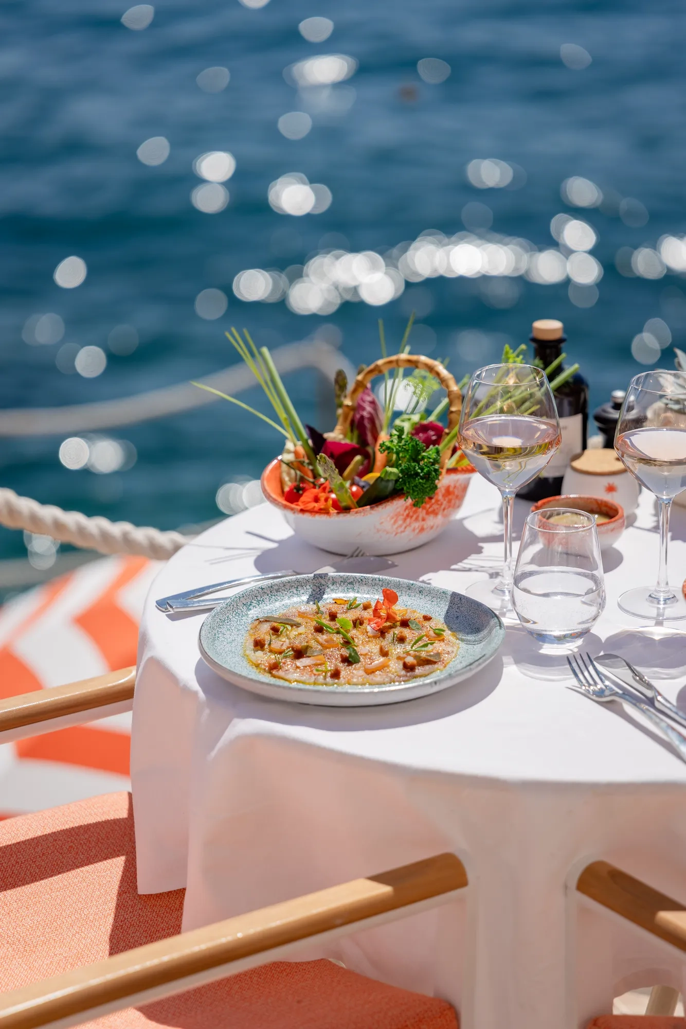 Seafood dish and salad served on a white table by the water, with wine glasses and sunlight reflecting on the sea.