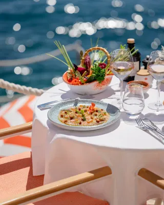 Plat de fruits de mer et salade servis sur une table blanche au bord de l’eau, avec verres de vin et reflets du soleil sur la mer.