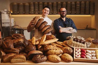 Two bakers smiling behind a counter piled with artisan breads, one holding loaves, with pastries and baked goods displayed in a warm bakery setting