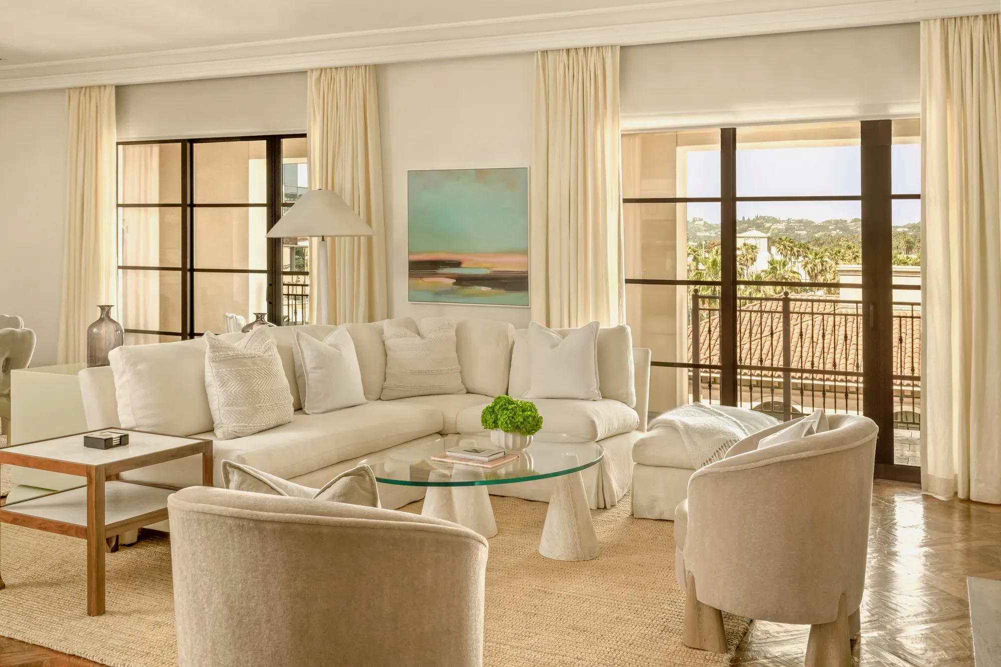 Elegant living room with cream seating, glass coffee table, tall drapes, and balcony views through large black-framed doors.
