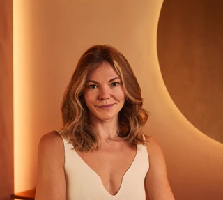 Woman in a cream sleeveless dress standing with arms folded, smiling at camera in a warmly lit interior with curved wall detail.