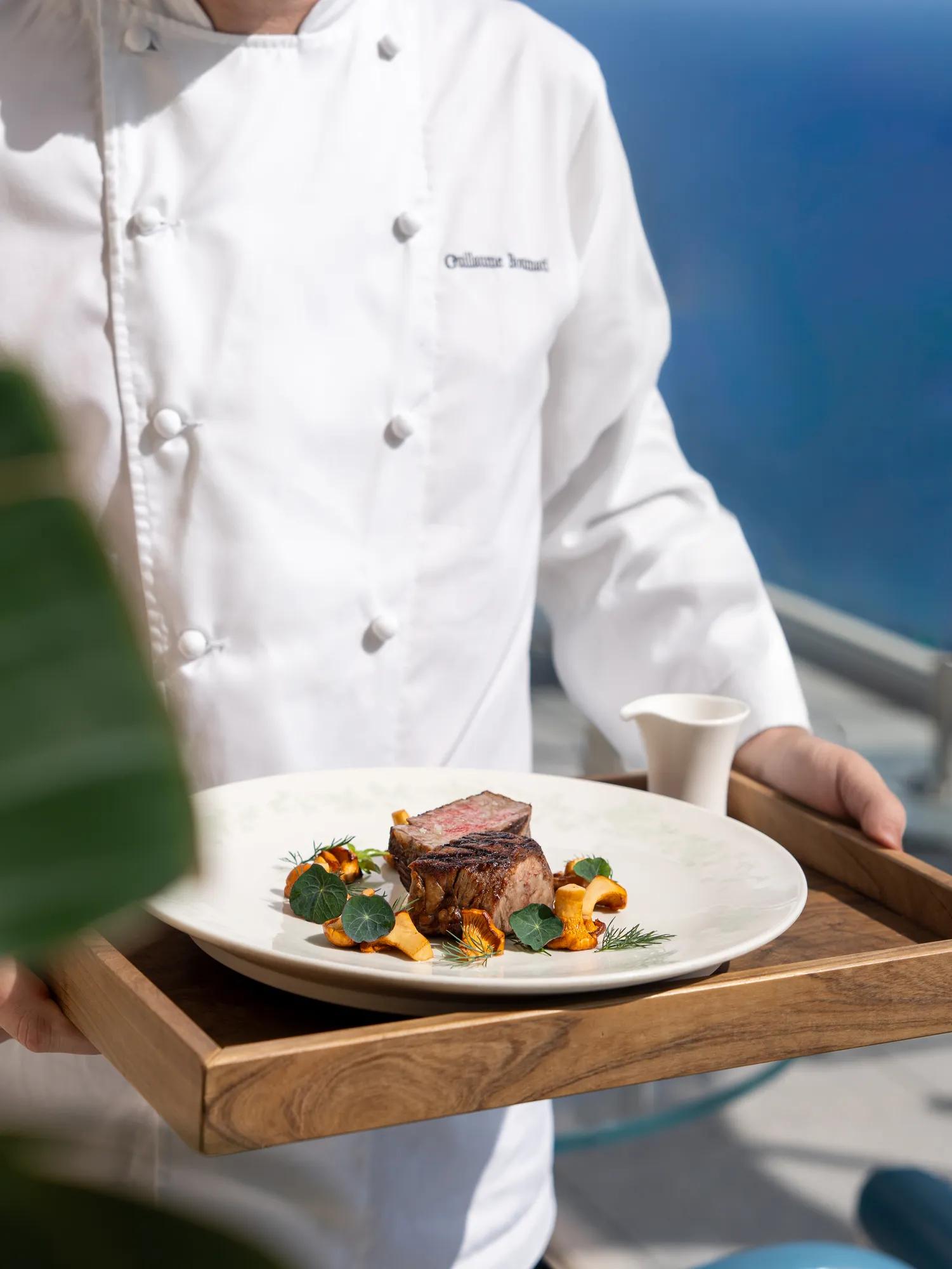 Chef en uniforme blanc présentant un plat gastronomique de viande saisie avec légumes de saison sur un plateau en bois, avec la mer en arrière-plan.