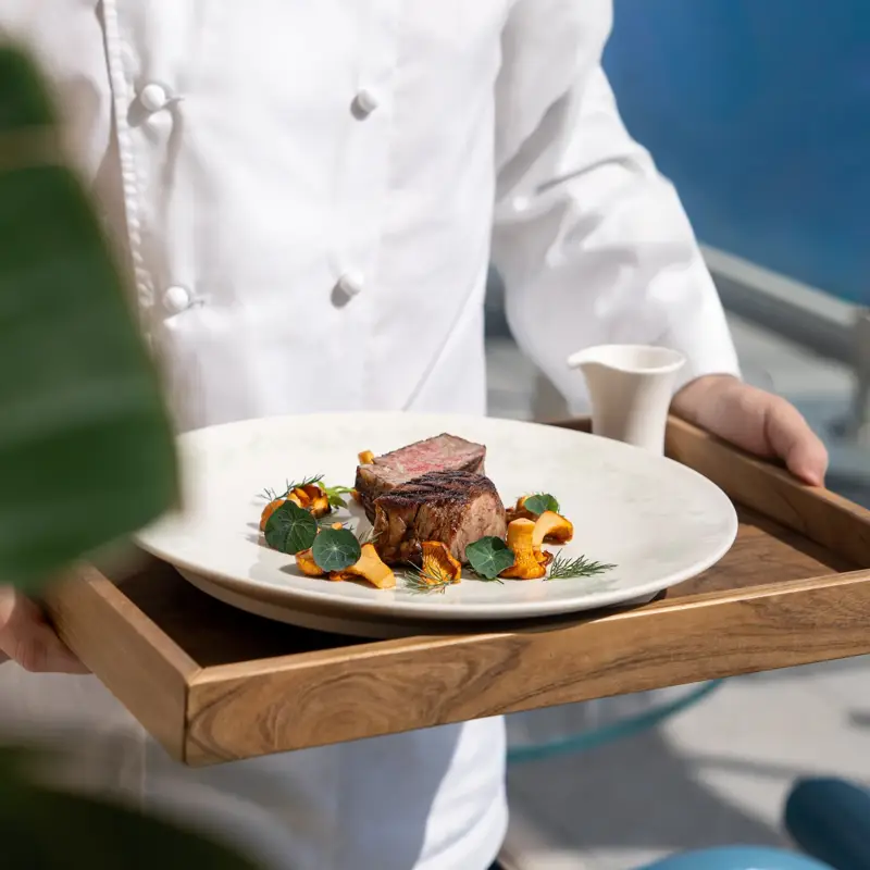Chef en uniforme blanc présentant un plat gastronomique de viande saisie avec légumes de saison sur un plateau en bois, avec la mer en arrière-plan.