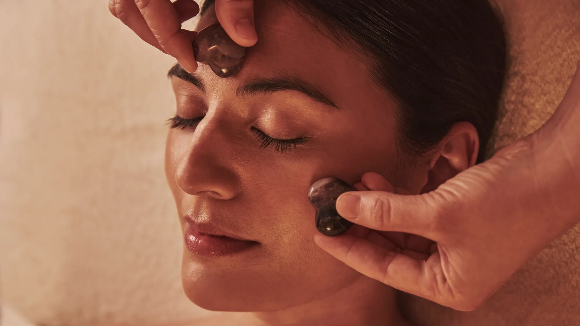 Person receiving a facial massage with smooth stones applied to the forehead and cheek while lying relaxed.