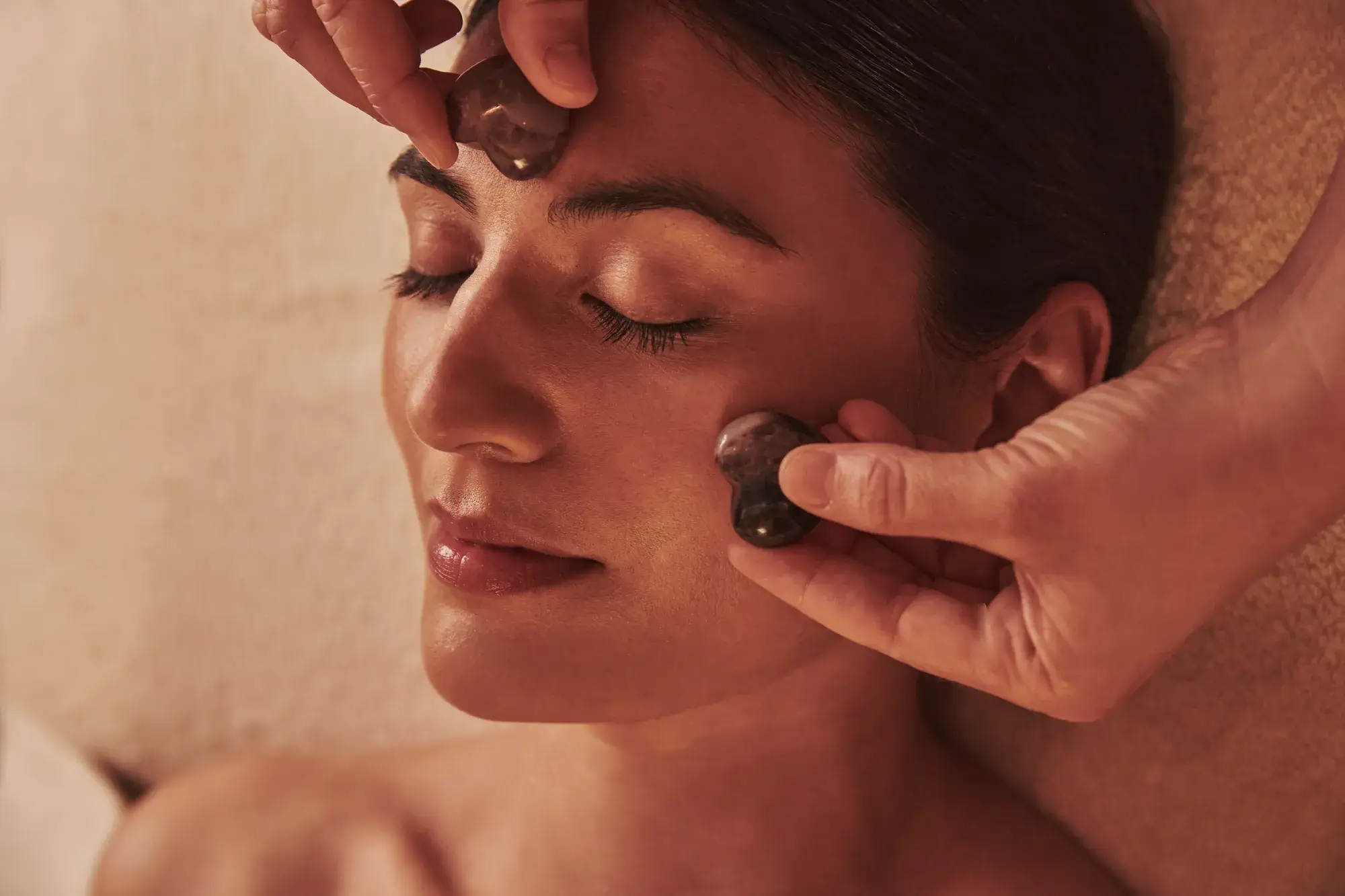 Person receiving a facial massage with smooth stones applied to the forehead and cheek while lying relaxed.
