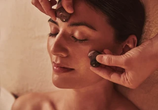 Person receiving a facial massage with smooth stones applied to the forehead and cheek while lying relaxed.