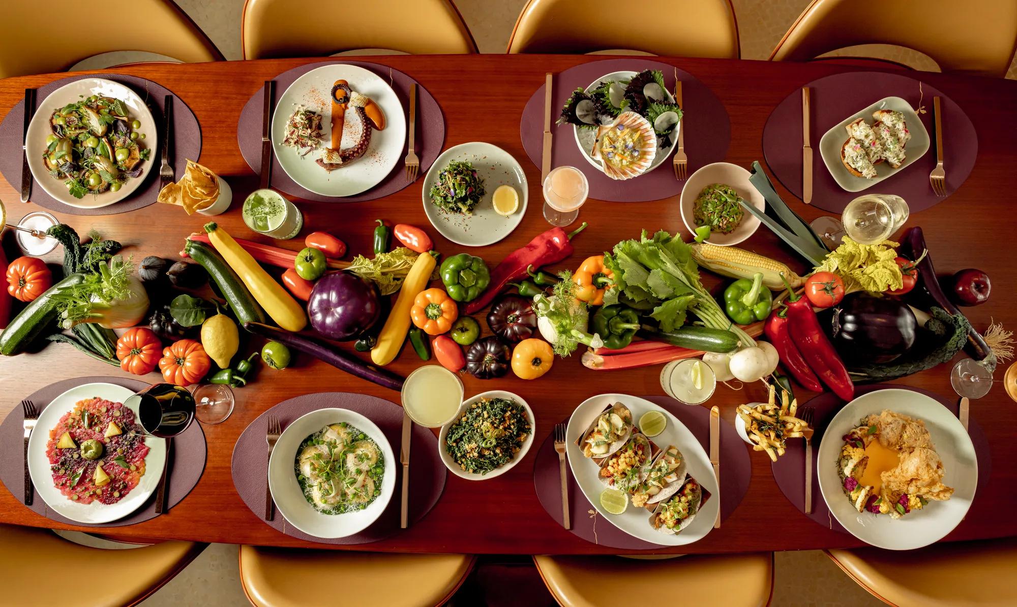 A dining table arranged with gourmet dishes and cocktails, surrounded by golden chairs, with a vibrant display of fresh vegetables including peppers, aubergines, courgettes, tomatoes and corn running down the centre as decoration.