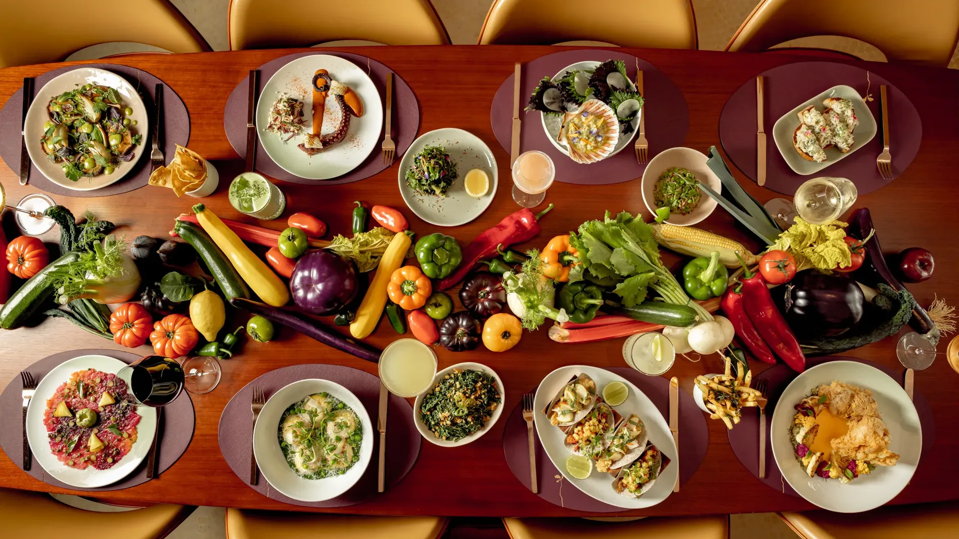 A dining table arranged with gourmet dishes and cocktails, surrounded by golden chairs, with a vibrant display of fresh vegetables including peppers, aubergines, courgettes, tomatoes and corn running down the centre as decoration.