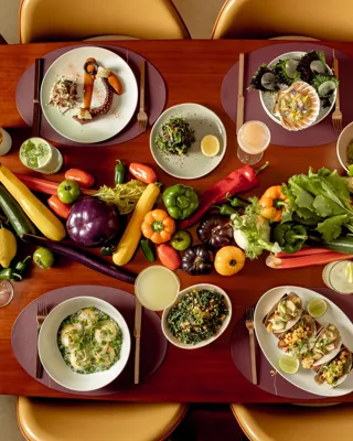 A dining table arranged with gourmet dishes and cocktails, surrounded by golden chairs, with a vibrant display of fresh vegetables including peppers, aubergines, courgettes, tomatoes and corn running down the centre as decoration.