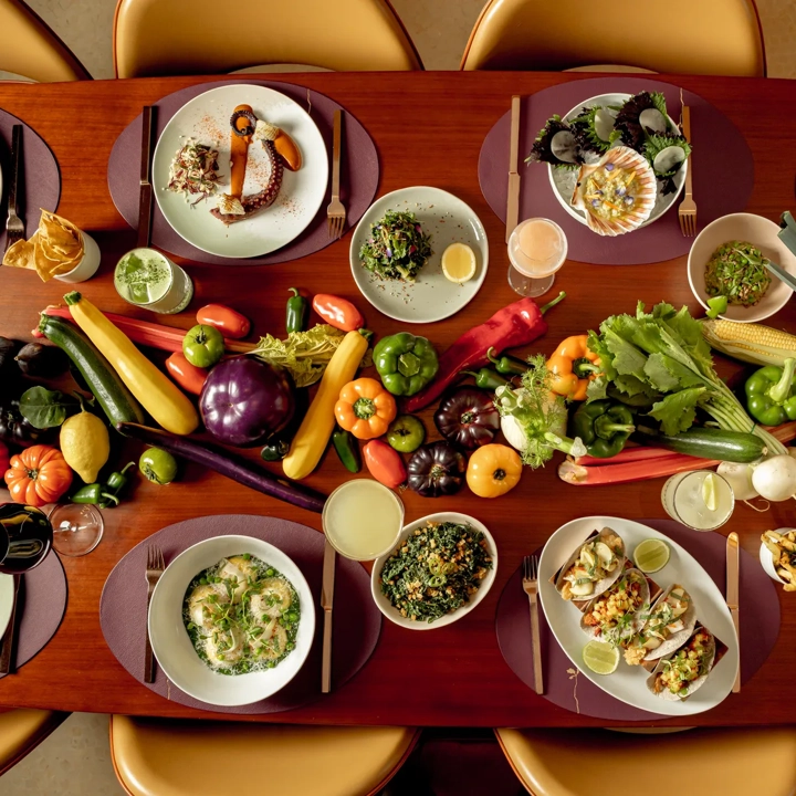 A dining table arranged with gourmet dishes and cocktails, surrounded by golden chairs, with a vibrant display of fresh vegetables including peppers, aubergines, courgettes, tomatoes and corn running down the centre as decoration.