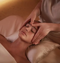 A person lies on a massage table with eyes closed while a therapist gently massages their forehead and temples using both hands in a softly lit spa setting.
