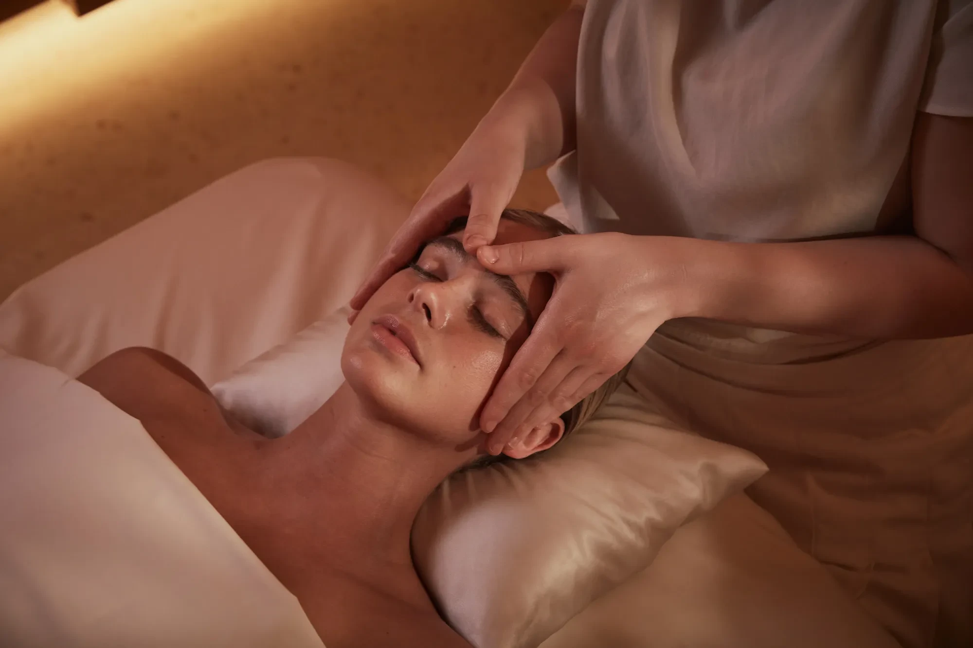 A person lies on a massage table with eyes closed while a therapist gently massages their forehead and temples using both hands in a softly lit spa setting.