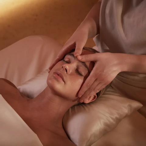 A person lies on a massage table with eyes closed while a therapist gently massages their forehead and temples using both hands in a softly lit spa setting.